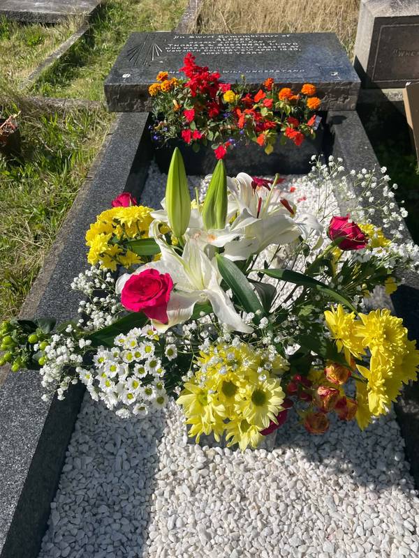 A kerbed grave of white gravel with a planted trough and an arrangement of lilies, yellow chrysanthemums, roses and gypsophila