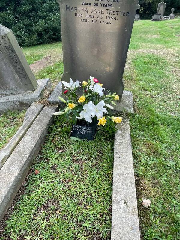 A weathered sandstone headstone on a kerbed grave, with white lilies and yellow roses in a black vase